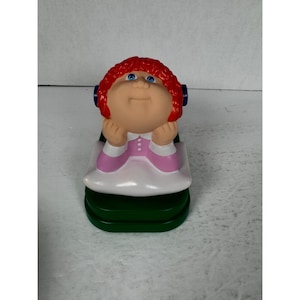 May include: A Cabbage Patch Kids figurine featuring a doll with red hair, blue eyes, and a pink and white dress. The doll is resting on a green base. The figurine is a collectible item.