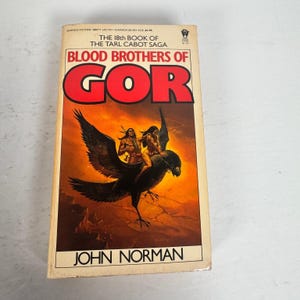 May include: A vintage paperback book titled "Blood Brothers of Gor" by John Norman. The cover features a fantasy illustration of two figures riding a large black bird against a fiery orange background. The book is part of "The Tarl Cabot Saga."