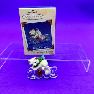 May include: A Hallmark Keepsake Ornament from the Collector's Series, featuring a white polar bear wearing a green scarf and blue hat, perched on the year "2003". The ornament has a red cardinal and the words "Cool Decade". The box is visible.