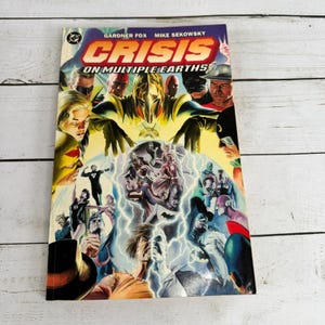Puede incluir: Un cómic vintage titulado "Crisis on Multiple Earths" de Gardner Fox y Mike Sekowsky. La portada presenta un collage de ilustraciones coloridas de superhéroes con el logo de DC Comics en la esquina superior izquierda. El fondo es amarillo claro con varios personajes.