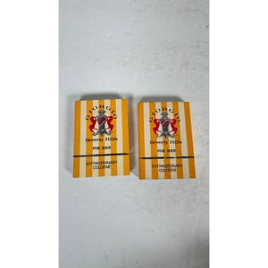 May include: Two rectangular cologne boxes featuring a vertical yellow and white striped pattern. The text reads Giorgio Beverly Hills, For, and Extraordinary Cologne, accompanied by a central crest logo with two red figures on a dark background.