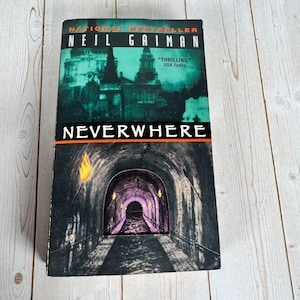 Pode incluir: Um livro de bolso intitulado "NEVERWHERE" de Neil Gaiman. A capa apresenta uma ilustração escura de um túnel com uma extremidade brilhante e uma paisagem urbana. O livro é um "NATIONAL BESTSELLER".