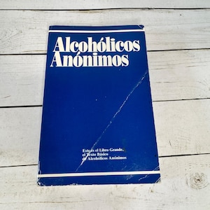 Op de afbeelding: Een blauw boek met de titel "Alcoholicos Anónimos" in witte letters. De omslag bevat ook de tekst "Este es el Libro Grande, el Texto Básico de Alcohólicos Anónimos."