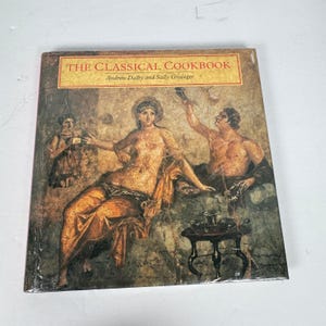 Puede incluir: Un libro de cocina de tapa dura titulado "The Classical Cookbook" con una portada que presenta una pintura clásica de figuras en un entorno romano. El título está en letras doradas sobre una pancarta marrón. El libro está sellado en plástico.