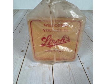 May include: A vintage Stroh's Country Inn sign, encased in clear plastic. The sign is rectangular with a light brown background and red lettering. The text reads "WELCOME YOU'RE IN Stroh's COUNTRY". The sign is on a light wood surface.