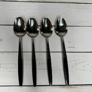 May include: Four stainless steel spoons with a decorative floral pattern on the handles and black handles. The spoons are arranged in a row on a white wooden surface.