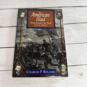 Puede incluir: Un libro de tapa dura titulado "An American Iliad: The Story of the Civil War" de Charles P. Roland. La portada presenta una ilustración en blanco y negro de soldados y un cañón, con retratos de figuras históricas. El libro tiene un esquema de color azul y marrón.