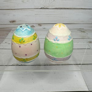 May include: Two decorative Easter egg-shaped salt and pepper shakers. One egg is light blue, yellow, and pink with purple polka dots. The other is green, pink, and white with blue floral accents. The shakers are on a clear surface.