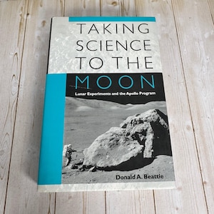 May include: A hardcover book titled Taking Science to the Moon: Lunar Experiments and the Apollo Program by Donald A. Beattie. The cover features a black and white photograph of a lunar landscape with a large rock and an astronaut.