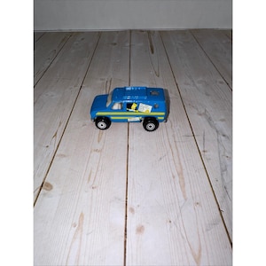 May include: A blue toy van with yellow and white accents. The van has black wheels and a yellow stripe along the side. There is a small sticker on the side of the van. The van is sitting on a wooden surface.