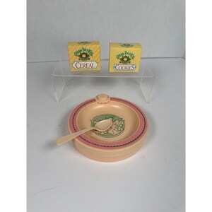 May include: A vintage Cabbage Patch Kids toy set. It includes a peach-coloured bowl with a pink rim, a spoon, and two small boxes labelled "Cereal" and "Cookies". The boxes are yellow with green and blue text.