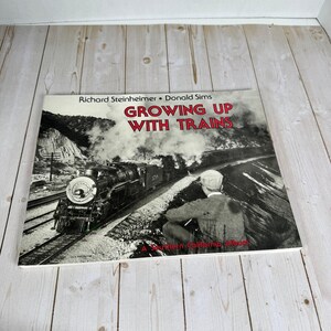 May include: A hardback book titled Growing Up With Trains by Richard Steinheimer and Donald Sims. The cover features a black and white photograph of a steam locomotive on tracks with a person in a cap sitting in the foreground.