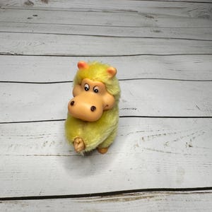 May include: A vintage plush toy hippo with a yellow body and a tan face. The hippo has black eyes and small pink ears. The toy is standing on a white wooden surface.