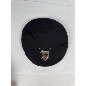 May include: Black, round hat with a Highlander emblem. The emblem features a shield with a castle, crossed swords, and the word "Highlander". The hat is made of a smooth, dark fabric.