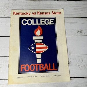 Puede incluir: Programa de fútbol universitario vintage para el partido Kentucky contra Kansas State. La portada presenta un gráfico de antorcha y fútbol en rojo, blanco y azul sobre un fondo azul oscuro. El texto dice «FÚTBOL UNIVERSITARIO».