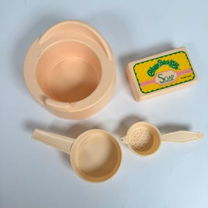 May include: A collection of vintage Cabbage Patch Kids bath toys in a light peach color. Includes a small potty, a soap bar with the word "SOAP", a small ladle, and a strainer.