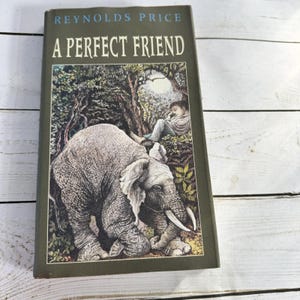 May include: A hardcover book titled A Perfect Friend by Reynolds Price with a dark olive green cover. The front features an illustration of a large gray elephant in the foreground and a person sitting in a forest setting in the background.
