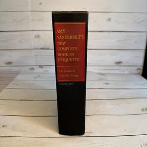 May include: A hardcover book titled "Amy Vanderbilt's New Complete Book of Etiquette." The book has a black spine with a red rectangular label containing the title and author's name. The book is a guide to gracious living, published by Doubleday.