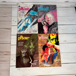 Może przedstawiać: Cztery komiksy w stylu vintage zatytułowane "The Prisoner" autorstwa Deana Mottera. Okładki zawierają ilustracje w różnych kolorach, w tym niebieskim, czerwonym i zielonym. Każda książka jest oznaczona literą A, B i C i ma metkę z ceną.