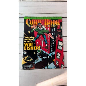 Puede incluir: Un cómic vintage titulado "COMIC BOOK PLACE" con una ilustración de portada de un hombre que lleva a una mujer, ambientada en un paisaje urbano. La portada presenta el texto "Working without a net... Will EISNER!" y es una revista para coleccionistas.