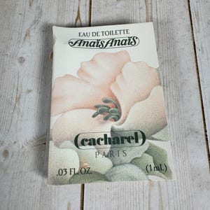 Könnte beinhalten: Eine rechteckige Probekarte für Anais Anais Eau de Toilette von Cacharel. Die weiße Karte zeigt eine große, sanft getönte Illustration einer blassrosa und grünen Blume. Der Text umfasst EAU DE TOILETTE, Anais Anais, cacharel, PARIS und (1mL).