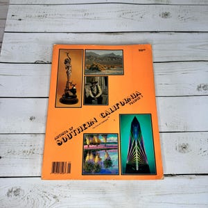Puede incluir: Un libro naranja titulado "Artistas del Sur de California Volumen 1" con varias imágenes de arte. La portada presenta una estatua, un paisaje, un hombre con sombrero y un diseño abstracto. El libro está sobre una superficie de madera blanca.