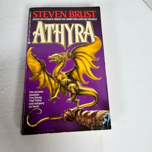 May include: A paperback book titled "Athyra" by Steven Brust. The cover features a golden dragon with outstretched wings against a purple background. Text includes the author's name and a tagline about the story's characters.