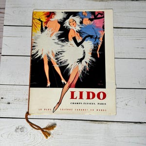 Puede incluir: Folleto de viaje vintage para el cabaret Lido en París. La portada presenta una ilustración de bailarinas con trajes de plumas blancas y azules. La palabra "LIDO" está en rojo, con el texto "Champs-Élysées, Paris" debajo.