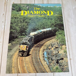Könnte beinhalten: Ein Vintage-Magazin mit dem Titel The Diamond Volume 7 Number 1, das ein Foto einer gelb-grauen Diesellokomotive zeigt, die einen mit Autos beladenen Güterzug auf einer kurvigen Strecke durch einen dichten Wald und felsiges Gelände zieht.
