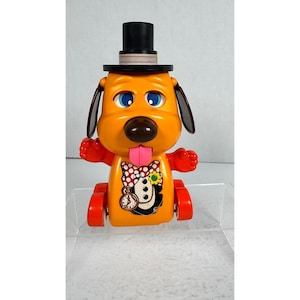 May include: A vintage orange toy dog with a black top hat, large blue eyes, and a pink tongue. The dog has red arms and wheels, and a decorative bow tie with a clock design on its chest. A classic toy.