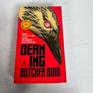 May include: A paperback book titled "Butcher Bird" by Dean Ing. The cover is predominantly red with a gold and black illustration of a bird's head. The text includes "Finally in Paperback" and "The Ransom of Black Stealth One".