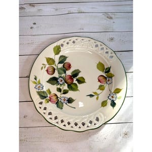 Peut inclure: Une assiette en céramique blanche avec une bordure ajourée décorative et un bord vert. L'assiette est ornée d'un motif floral avec des fruits rouges et violets, des feuilles vertes et de petites fleurs bleues. L'assiette est sur une surface en bois blanc.