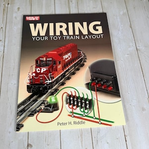 May include: A softcover book titled Wiring Your Toy Train Layout by Peter H. Riddle. The cover features a red CP Rail toy locomotive on tracks, a black electrical terminal block, and a diagram showing red and green wiring connections.