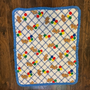 May include: A white baby blanket with a blue grid pattern and a blue ruffled border. The blanket features brown teddy bears and colorful balloons. The blanket measures approximately 30 inches by 30 inches.