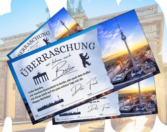 May include: A blue and white certificate with the text "Überraschung" and "wir fahren nach Berlin". The certificate is decorated with a silhouette of the Brandenburg Gate and the Berlin TV Tower. The text "Liebe Sandra, wir machen Kurzurlaub in Berlin, also pack dein Koffer. Ich freue mich auf ein paar schöne Tage mit dir. Happy Birthday!" is printed on the certificate. The certificate is signed "Dein Frank".