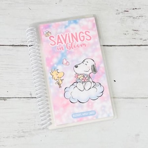 May include: A spiral-bound planner with a cover featuring Snoopy on a cloud, with Woodstock and a butterfly. The cover has the text "SAVINGS in bloom" in pink, with the text "BUDGET WITH CARLY" below the image, set against a pink and blue background.