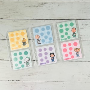 Puede incluir: Un conjunto de seis hojas de pegatinas cuadradas con personajes de Peanuts en ropa médica, incluyendo a Snoopy, Charlie Brown y Lucy. Cada hoja tiene un fondo de color con una cuadrícula de pegatinas circulares y el texto "SCRUBS WITH SNOOPY".