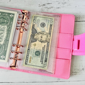 Könnte beinhalten: Ein pinkfarbenes Portemonnaie im Ordnerstil aus Kunstleder mit einem Ringbuch aus roségoldfarbenem Metall. Das Portemonnaie ist geöffnet und zeigt einen Zwanzig-Dollar-Schein in einer durchsichtigen Plastikhülle und einen Ein-Dollar-Schein.