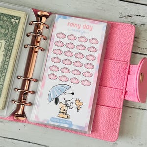 Könnte beinhalten: Ein rosa Planer mit einem roségoldenen Ordner und einem "Rainy Day Savings" Einsatz mit Snoopy und Woodstock unter einem Regenschirm. Der Einsatz hat rosa Wolkenillustrationen und den Text "Budget with Carly".