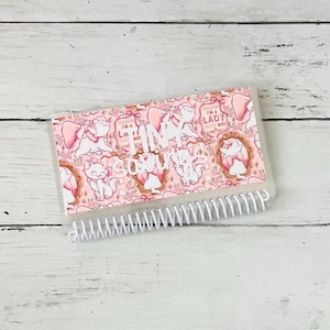 May include: A rectangular savings tracker with a white spiral binding at the bottom. The cover features a repeating pink pattern of white cartoon kittens with pink bows, floral accents, and the text "TINY savings" in white script lettering.