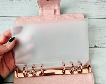 Könnte beinhalten: Ein hellrosa Planer mit roségoldfarbener Hardware. Der Planer ist geöffnet und zeigt eine transparente Kunststofftasche. Der Planer hat eine strukturierte lederartige Optik und einen Schnappverschluss. Die Metallringe sind sichtbar.