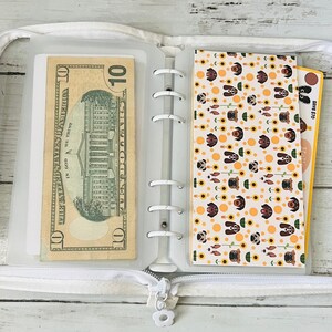 May include: A white zippered binder with a clear plastic pocket on the left side holding a $10 bill. The right side has a clear plastic pocket with a patterned sheet of paper featuring a repeating design of brown dogs and yellow sunflowers.