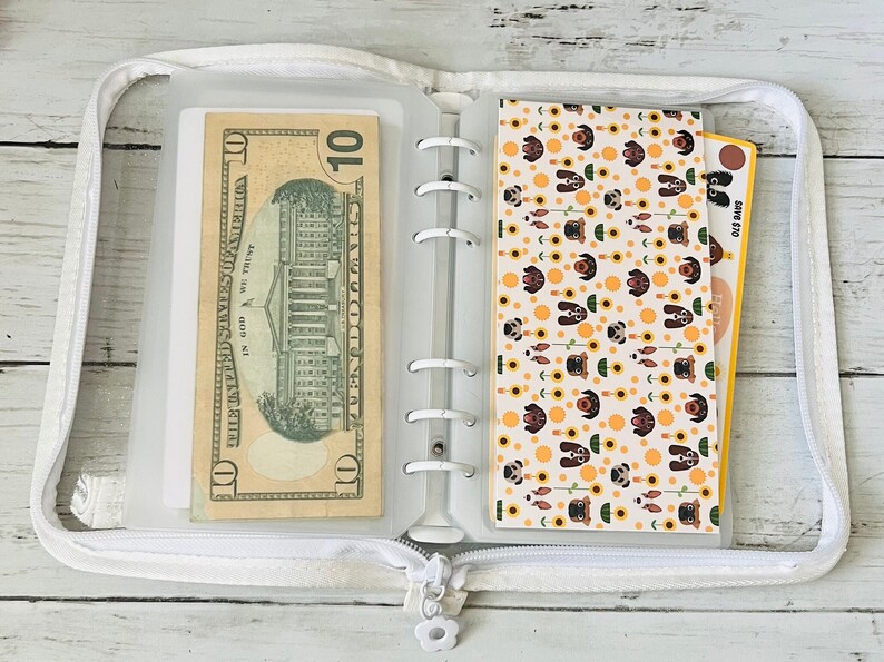 May include: A white zippered binder with a clear plastic pocket on the left side holding a $10 bill. The right side has a clear plastic pocket with a patterned sheet of paper featuring a repeating design of brown dogs and yellow sunflowers.