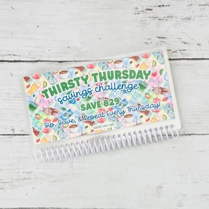 May include: A spiral-bound savings challenge notebook with the text "THIRSTY THURSDAY savings challenge SAVE 829". The cover features colorful illustrations of beverages and fruits. The notebook is white with a clear plastic cover.