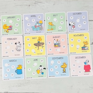 May include: A set of twelve square savings tracker cards featuring Peanuts characters for each month of the year. Each card displays a month name, illustrations of Snoopy or Woodstock, and circular spaces with numerical values for tracking savings.