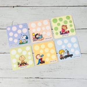 May include: Six square paper trackers featuring Peanuts characters with white circular fill-in spots and the text Budget with Carly. Designs include Snoopy listening to music, Lucy reading, Sally exercising, Charlie Brown meditating, Linus drinking, and Schroeder playing piano.