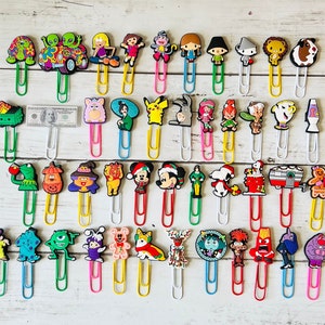 May include: A collection of 36 colorful cartoon character bookmark clips. The clips are made of plastic and feature a variety of characters, including a turtle, a monster, a dog, a cat, a reindeer, a snowman, a penguin, a unicorn, a dragon, a mermaid, a fairy, a princess, a prince, a pirate, a cowboy, a superhero, a villain, a robot, a dinosaur, a spaceship, a car, a truck, a train, a plane, a boat, a house, a tree, a flower, a sun, a moon, a star, a heart, and a rainbow.