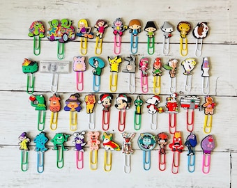May include: A collection of 36 colorful cartoon character bookmark clips. The clips are made of plastic and feature a variety of characters, including a turtle, a monster, a dog, a cat, a reindeer, a snowman, a penguin, a unicorn, a dragon, a mermaid, a fairy, a princess, a prince, a pirate, a cowboy, a superhero, a villain, a robot, a dinosaur, a spaceship, a car, a truck, a train, a plane, a boat, a house, a tree, a flower, a sun, a moon, a star, a heart, and a rainbow.