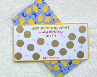 May include: A white card with a gold background and the text "When Life Gives You Lemons Savings Challenge Save £75" with 15 gold circles that can be scratched off. The card is on top of a blue and yellow patterned paper with lemons.