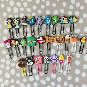 May include: A collection of 25 colorful, cartoon-themed metal bookmark clips. The clips feature characters like princesses, care bears, ghosts, and a variety of other fun designs.